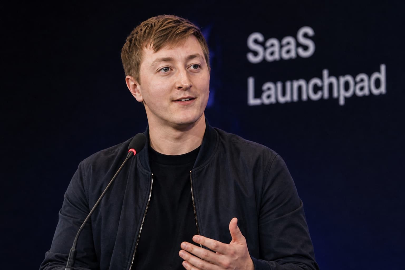 Moritz Zinecker speaking at a lectern microphone with blurred SaaS text in the background