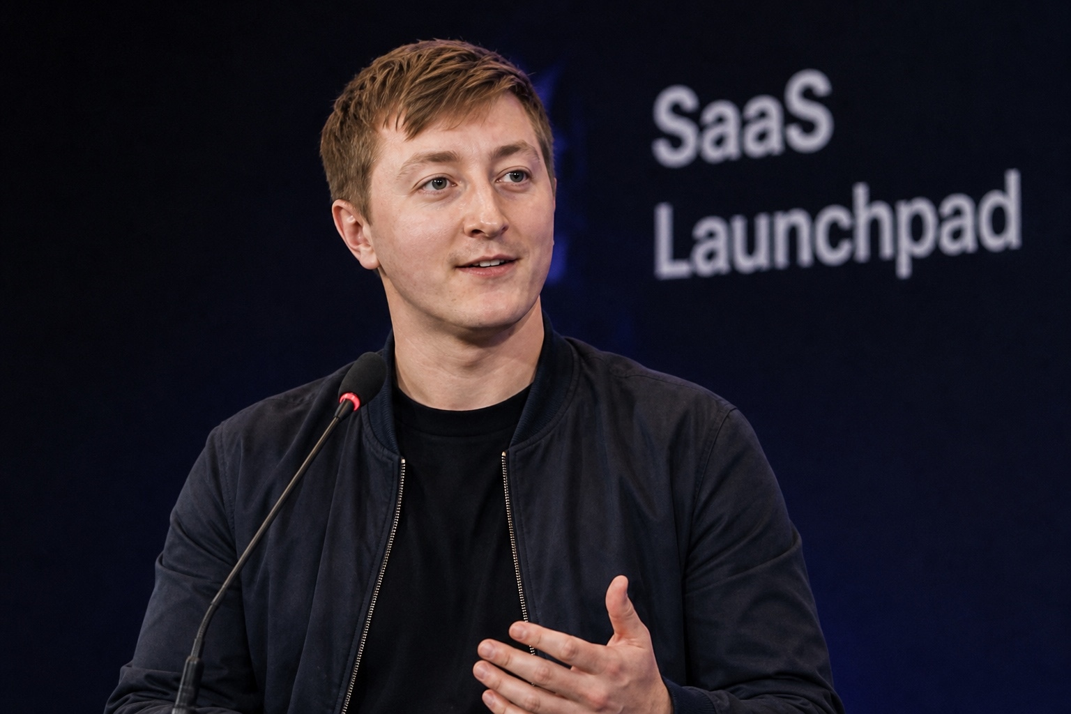 Moritz Zinecker speaking at a lectern microphone with blurred SaaS text in the background