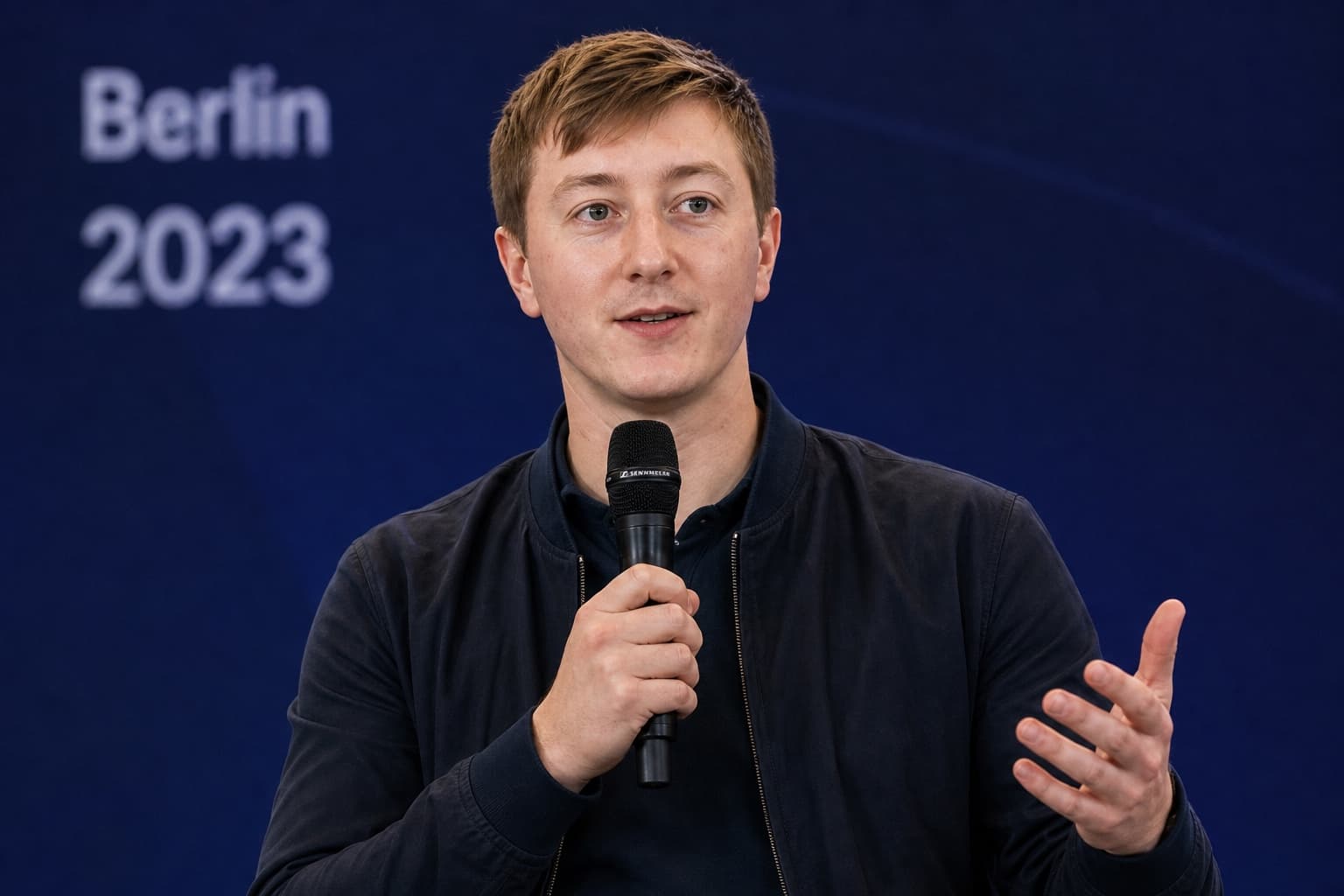 Moritz Zinecker speaking with a handheld microphone on a blue stage with Berlin 2023 visible in the background