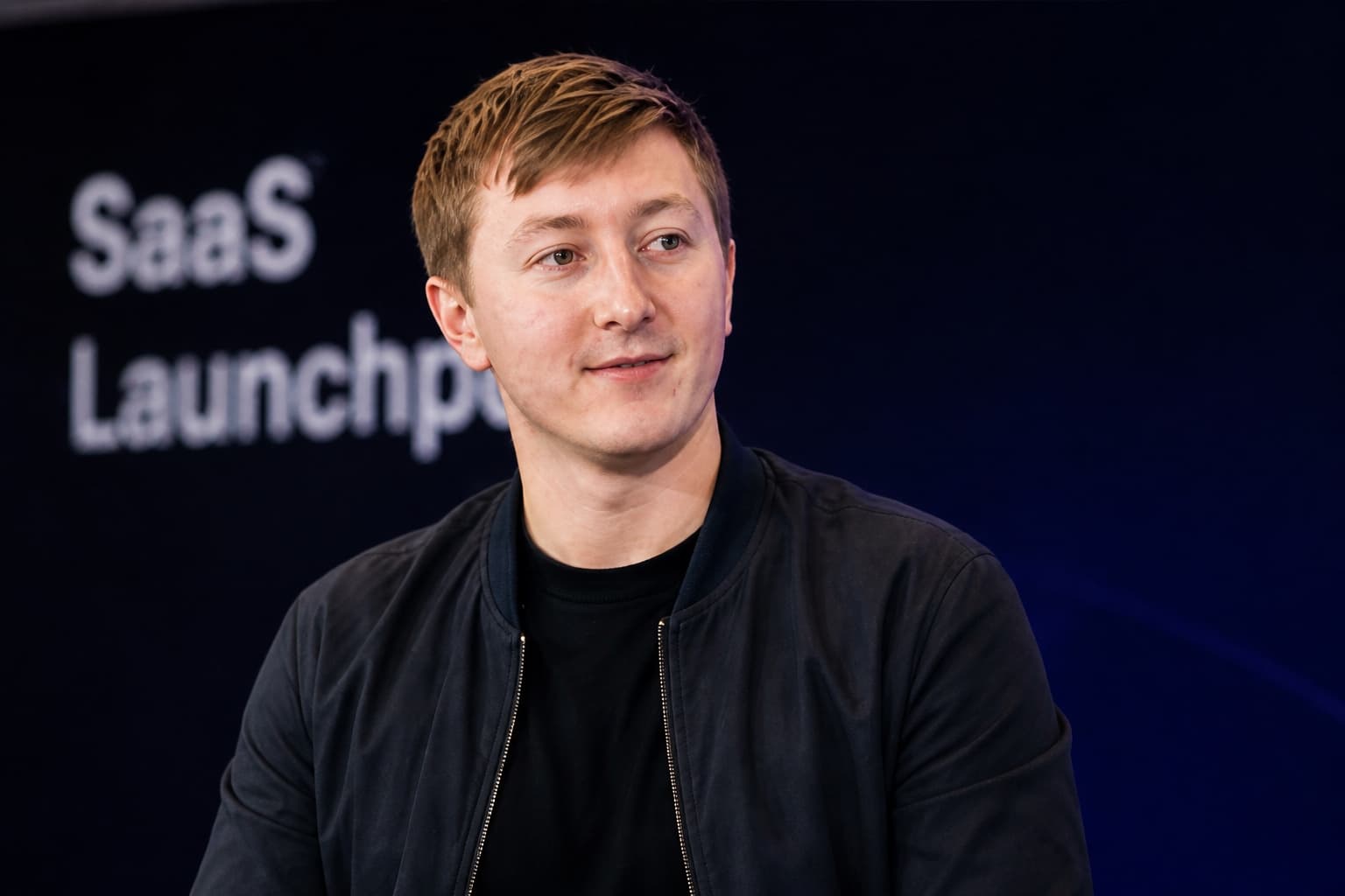 Moritz Zinecker seated on a dark stage with blurred SaaS text in the background